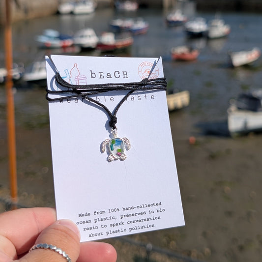 A handmade beach waste turtle pendant necklace on a seaweed and cotton cord, displayed on a white card held by a hand, with a blurred harbour and boats in the background.