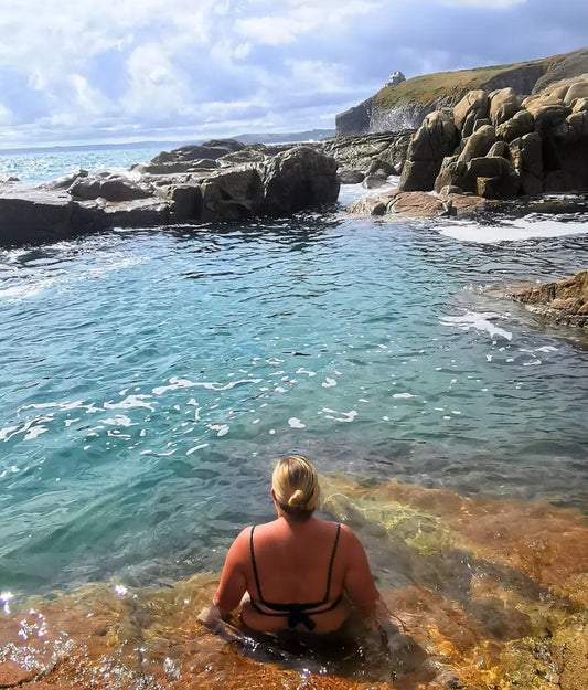 Rinsey tidal pool wild swimming. Woman sitting on the edge of Rinsey tidal pool in Cornwall.