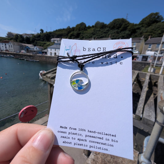 A hand holds a beach plastic wave pendant necklace on a white recycled display card outdoors by the seaside. The necklace has a wave shape silve pendant with blue, green, and yellow plastic pieces set inside with bioresin, attached to a black cord.