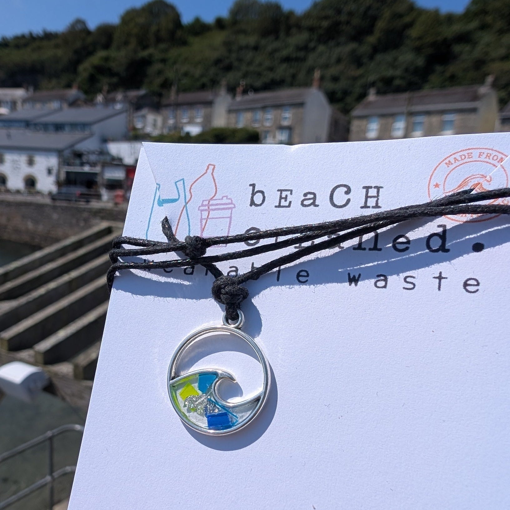 Sustainable ocean lover wave necklace gift. Wave shaped siver necklace with beach plastic pollution set within it on a black adjustable cord made from seaweed and cotton. 
