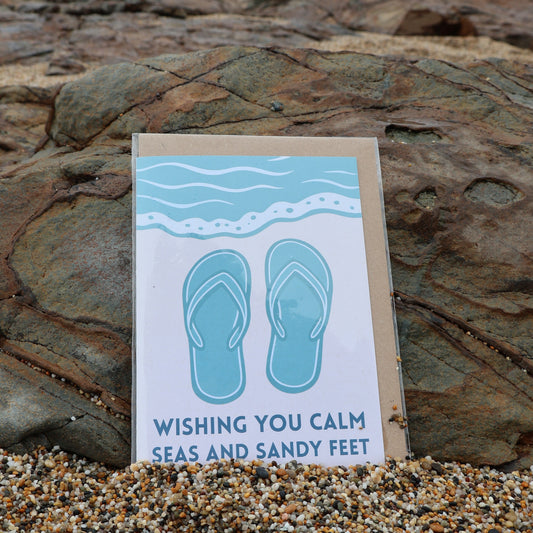 Card with flip-flop design and text 'Wishing you calm seas and sandy feet' on a rocky beach.