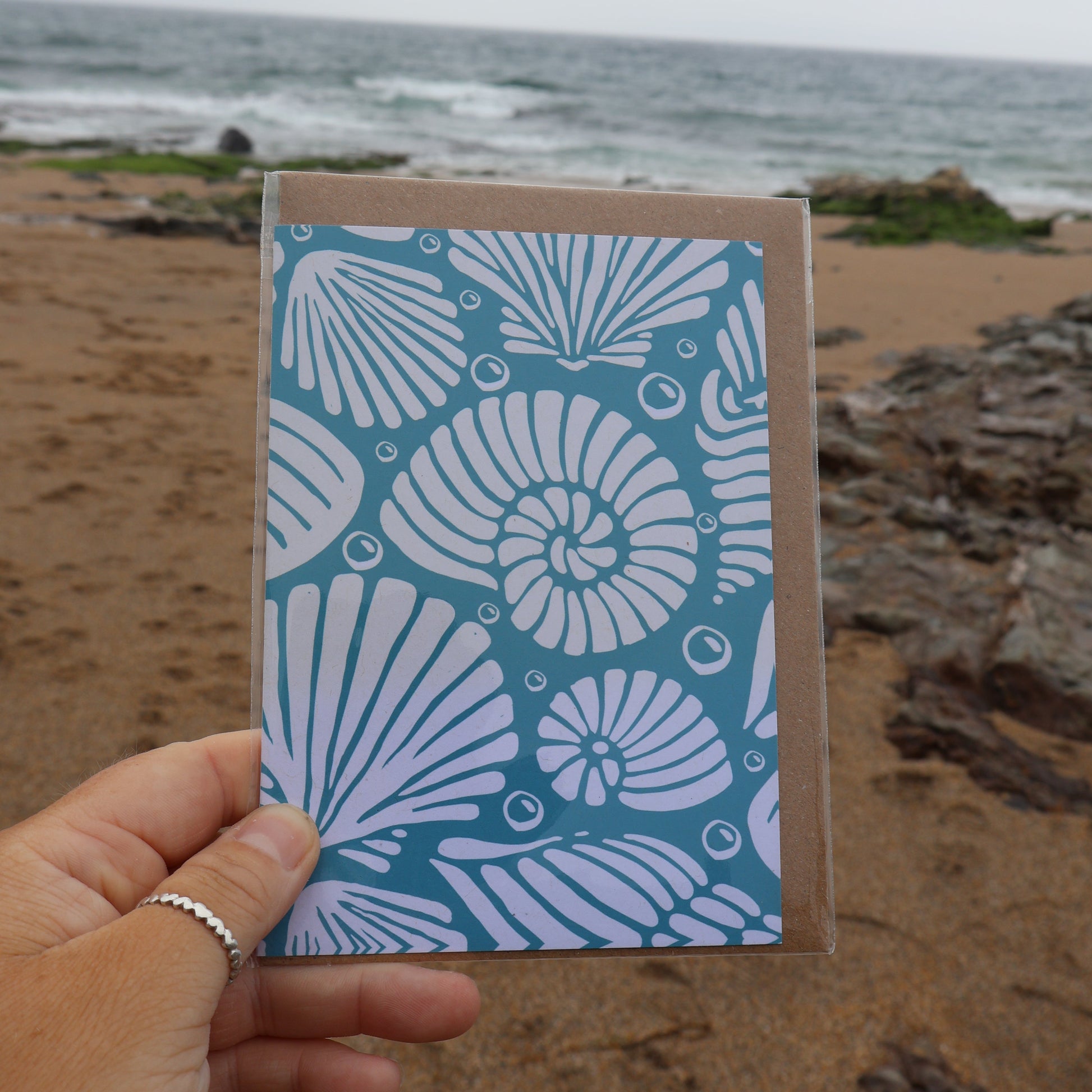 Blue recycled card with white shell pattern held by a hand on a beach