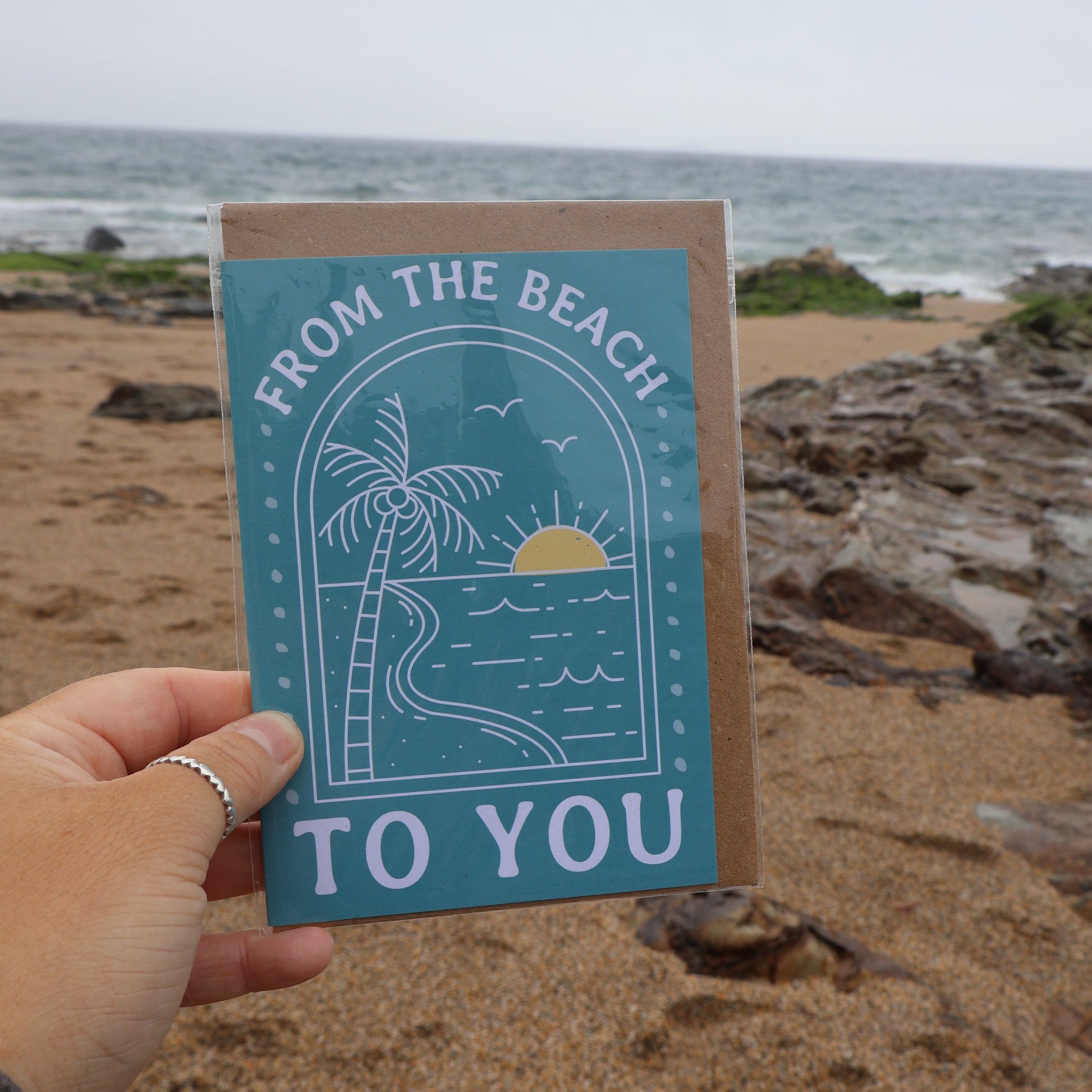 Card with 'From the Beach to You' text held by a hand on a beach.