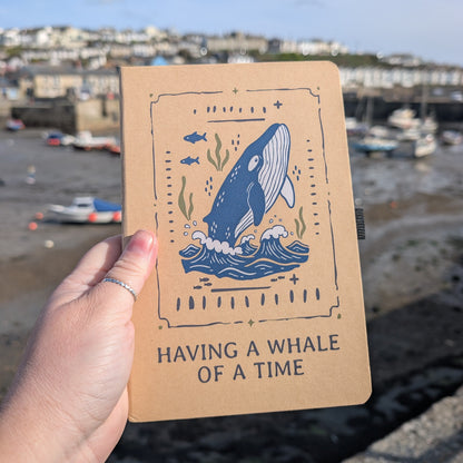 Notebook with whale illustration held by a hand, with a harbor background 