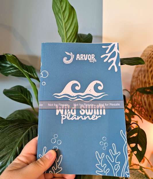 Blue wild swimming planner with white wave, seaweed and bubble designs held by a hand, with a plant in the background
