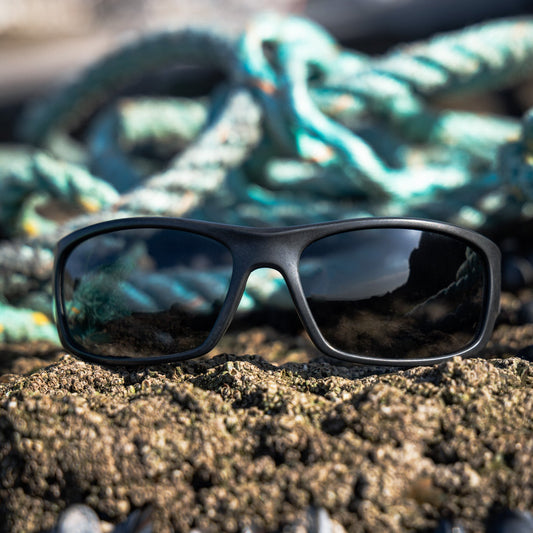 Black rectangular wrap-around recycled ocean waste sunglasses with dark lenses resting on black rocks, with fishing rope behind them.
