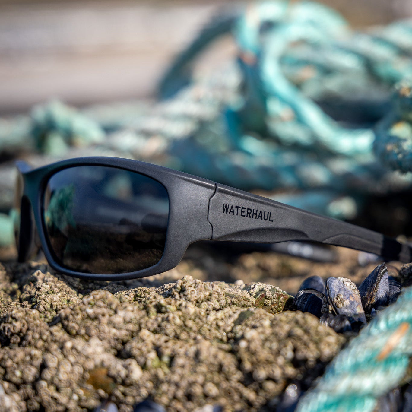 Waterhaul Mylor sunglasses resting on wet sand and mussels with reclaimed fishing gear in the background.