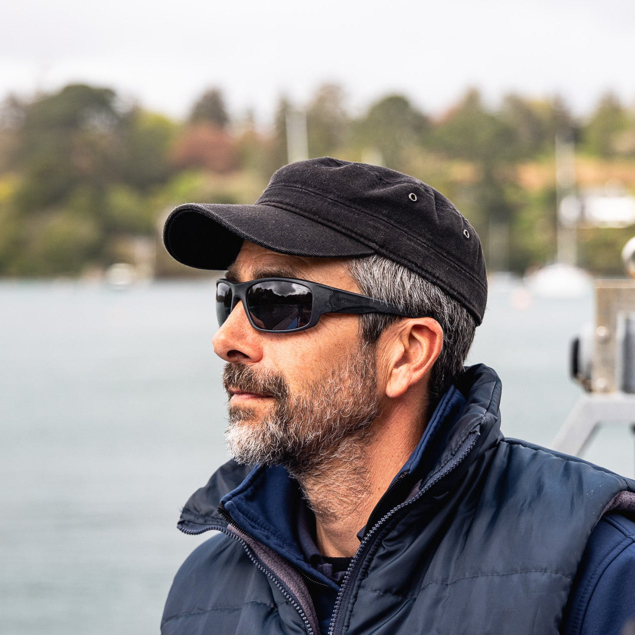 A man wearing wrap-around black sunglasses made from ocean waste sits on a boat with water and a tree-lined shore in the background.