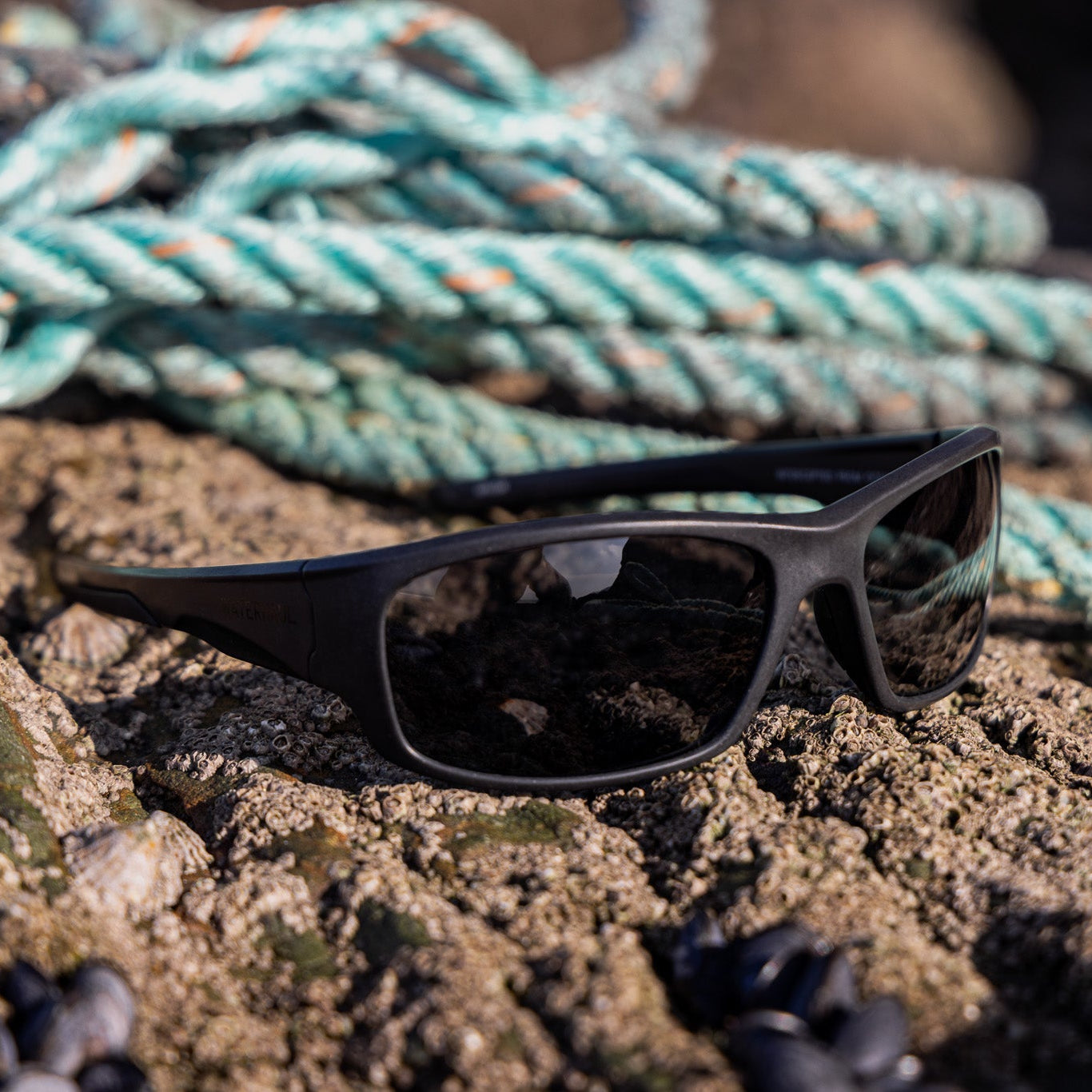 Black recycled plastic wrap-around sunglasses resting on wet sandy ground with a coiled fishing rope in the background.