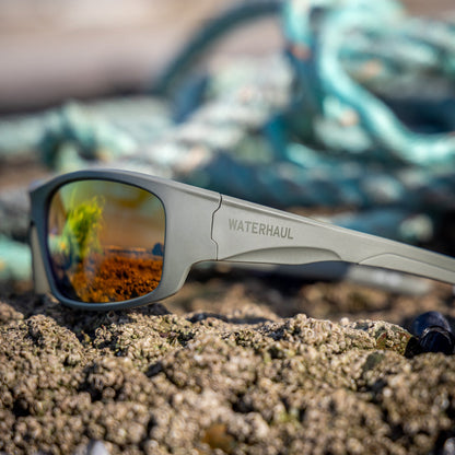 Gray-green Waterhaul sustainable sunglasses resting on a sandy beach with mussels in the foreground and a blurred fishing net in the background.