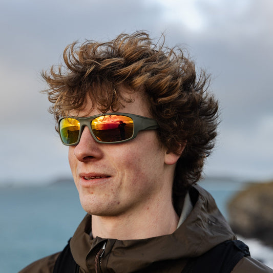 A man outdoors wearing recycled sustainable sunglasses with orange reflective mirrored lenses, by a coastal background.