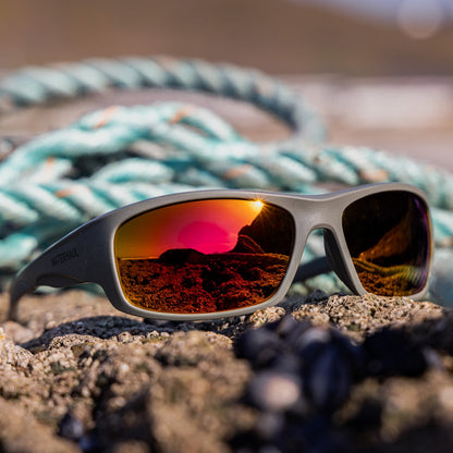Polarised wrap-around sunglasses with mirrored orange reflective lenses, resting on dark rocks in front of ghost nets collected from the beach. 