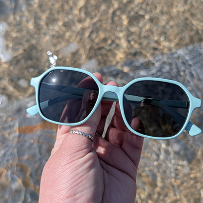 Hand holding light blue recycled ghost net sunglasses with a beach background