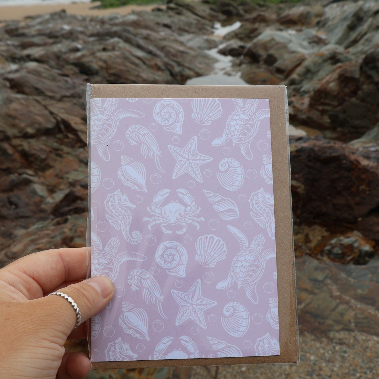 Hand holding a pink recycled card with a sea creature pattern at the beach