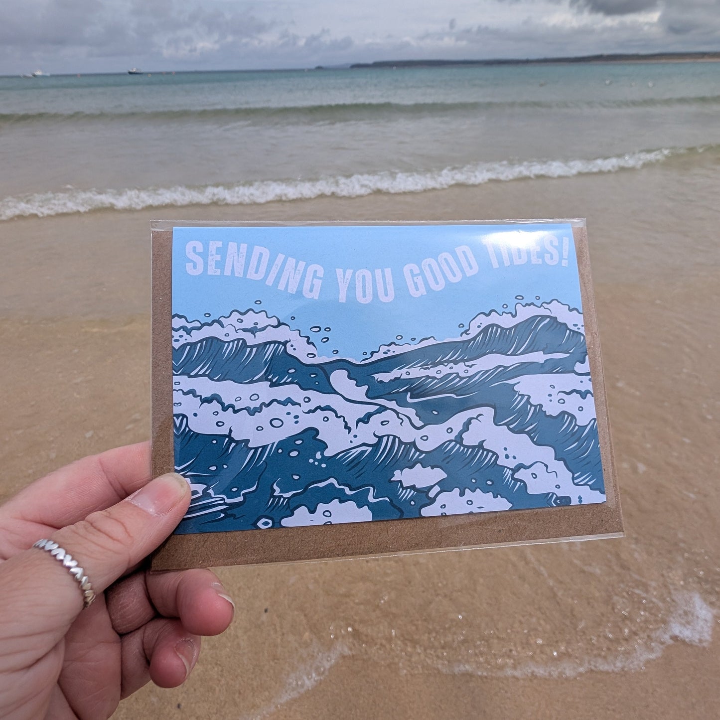 Hand holding a card with Sending You Good Tides Greetings Card ocean design on a beach
