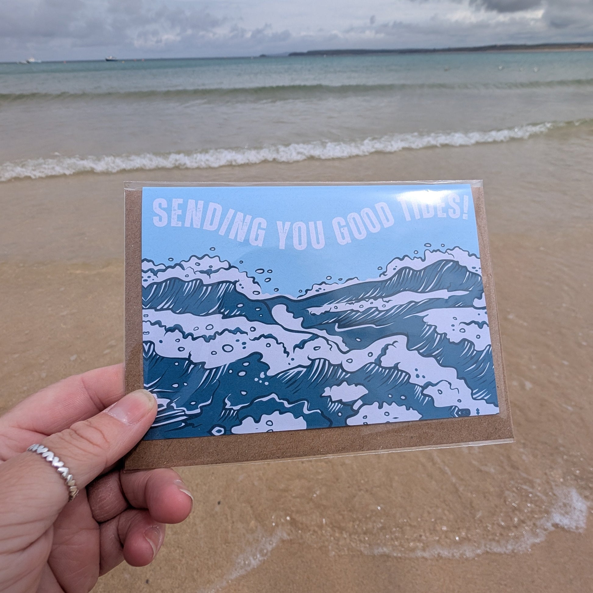 Hand holding a card with Sending You Good Tides Greetings Card ocean design on a beach