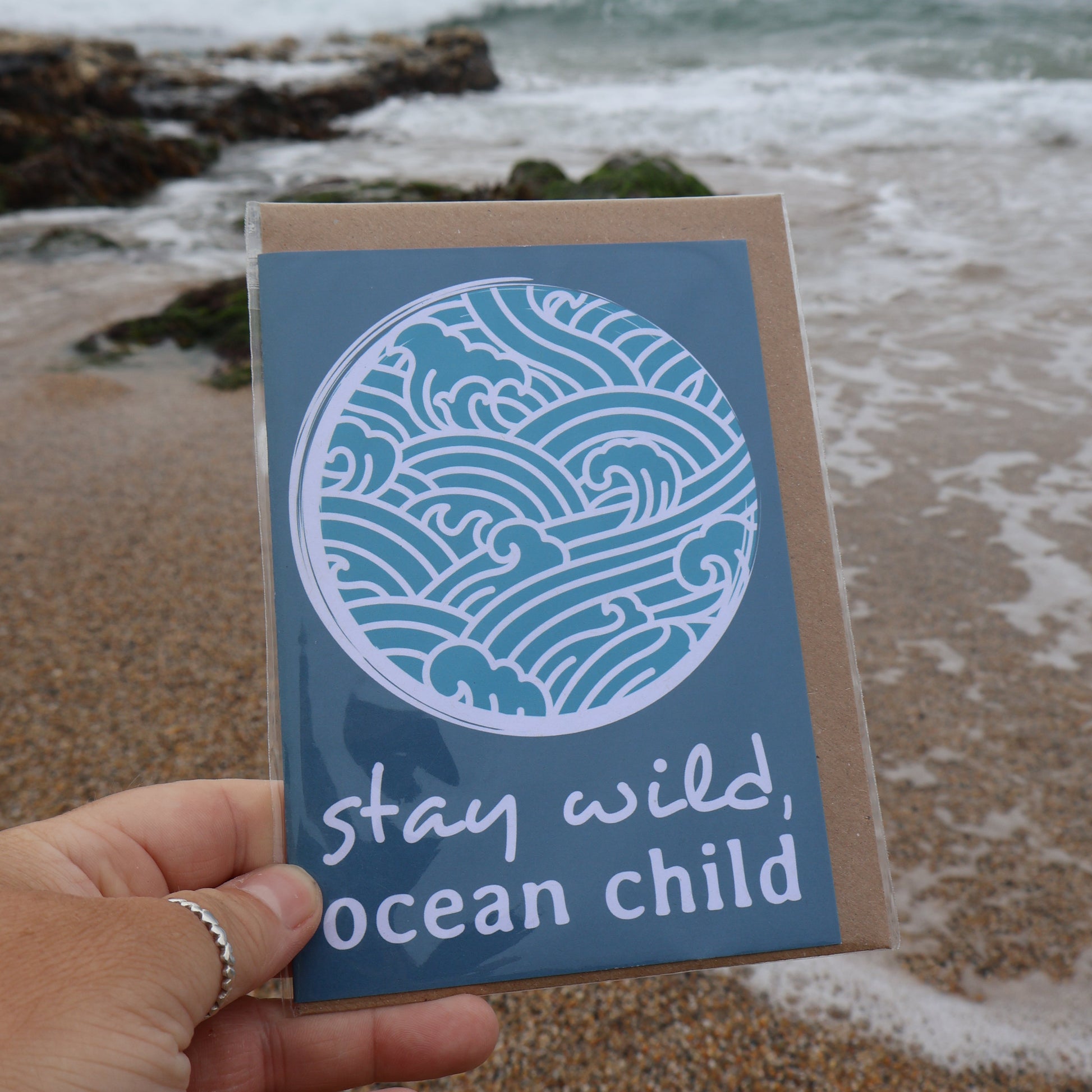 Hand holding a card with ocean-themed 'stay wild ocean child' design at the beach.