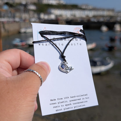 A hand holds a small seaweed cord necklace with a silver whale pendant which has beach plastic inside a bio resin. Sustainable necklace secured on a recycled card backing. 