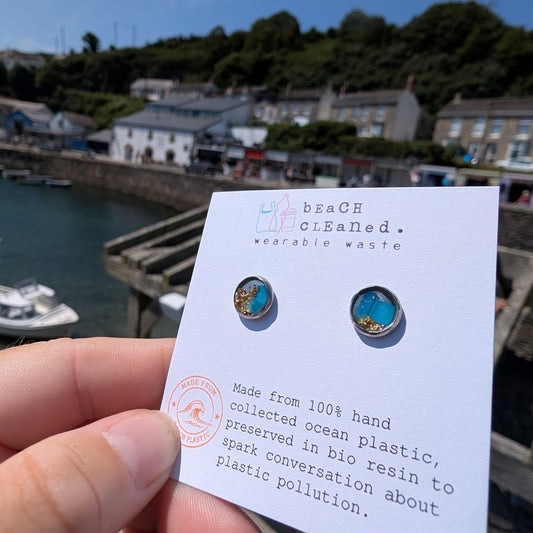 A hand holds a small white card displaying a pair of round stud earrings made from collected ocean plastic, with a harbor and buildings in the blurred background.