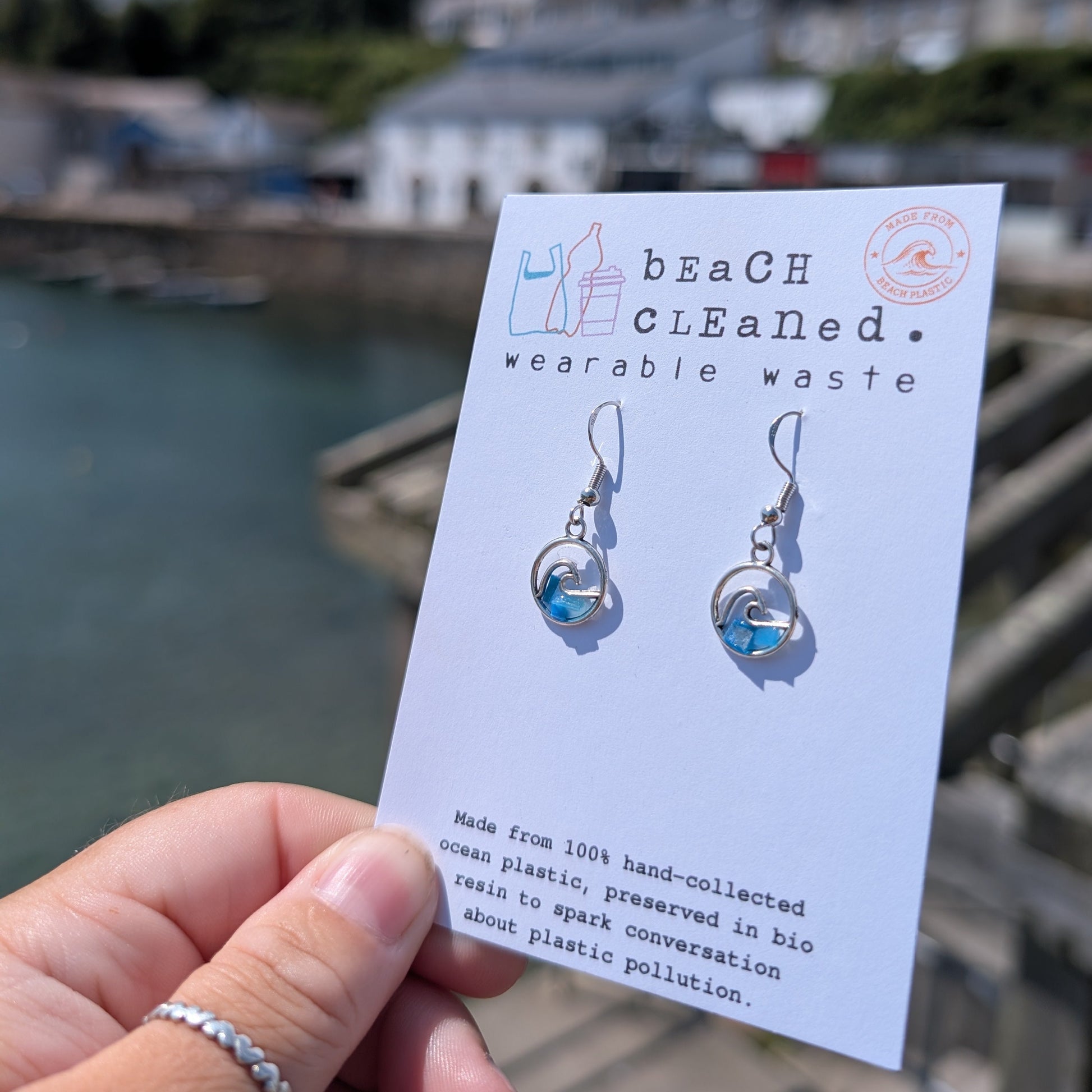 Sustainable sterling silver wave earrings made with beach plastic set in a bioresin. Displayed on recycled card beside the sea. 