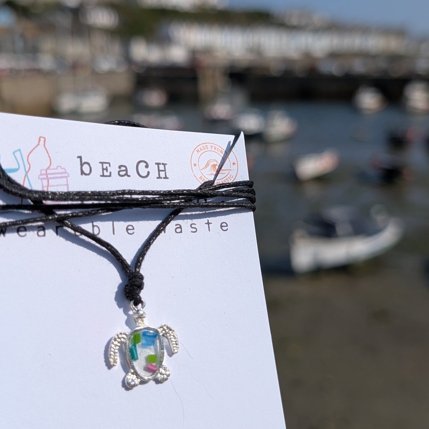 A silver turtle pendant necklace with a colorful iridescent center made from beach plastic, tied to a black cord and displayed on a white card printed with 'beach wearable taste'. The background is a blurred outdoor harbor scene with boats and buildings under a blue sky.