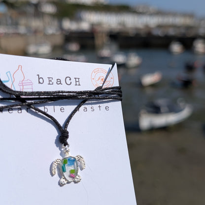 A silver turtle pendant necklace with a colorful iridescent center made from beach plastic, tied to a black cord and displayed on a white card printed with 'beach wearable taste'. The background is a blurred outdoor harbor scene with boats and buildings under a blue sky.