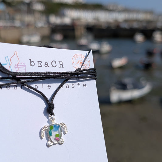 A silver turtle pendant necklace with a colorful iridescent center made from beach plastic, tied to a black cord and displayed on a white card printed with 'beach wearable taste'. The background is a blurred outdoor harbor scene with boats and buildings under a blue sky.