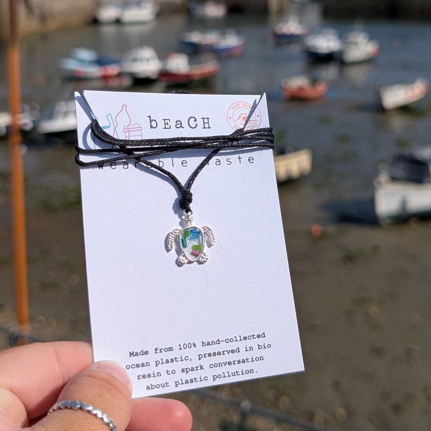 A handmade beach waste turtle pendant necklace on a seaweed and cotton cord, displayed on a white card held by a hand, with a blurred harbour and boats in the background.