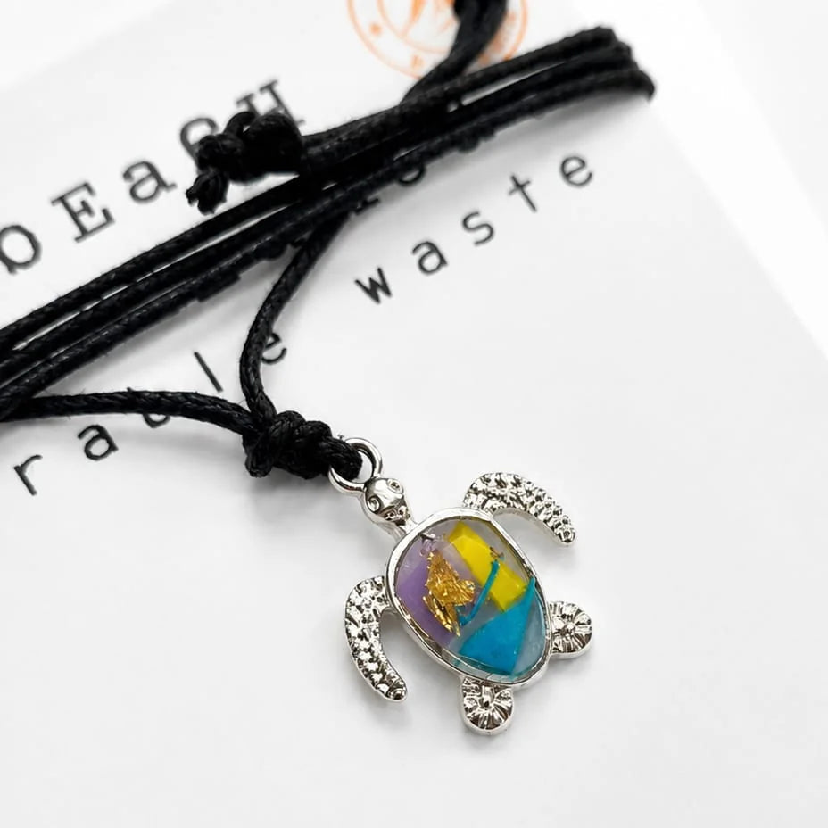 A silver sea turtle pendant necklace with a colorful shell made from plastic pollution threaded on a black cord and displayed on white recycled printed card.