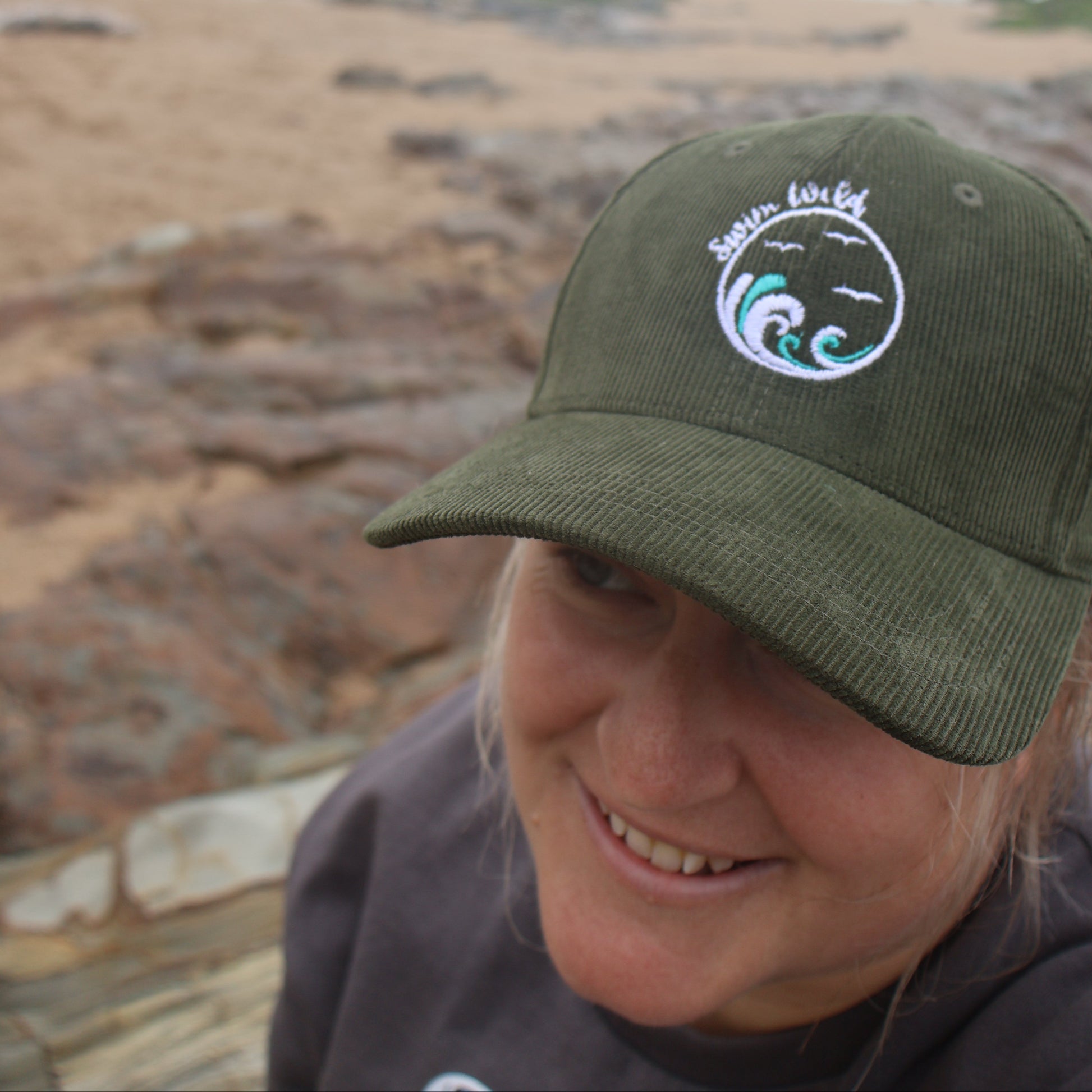 Person wearing a green cap with a wild swimming logo on a beach