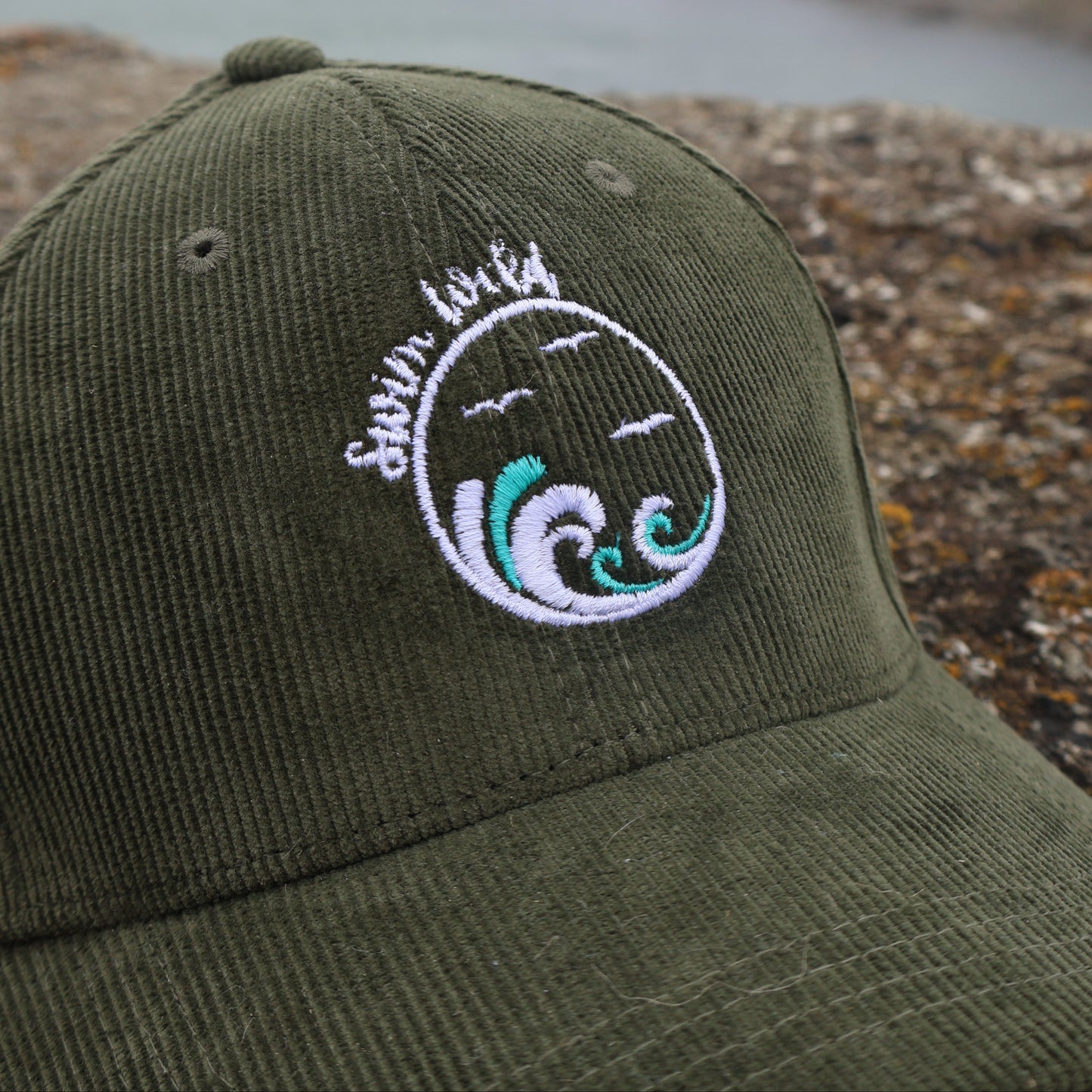 Green organic cotton cap with swim wild embroidered design on a rocky shoreline
