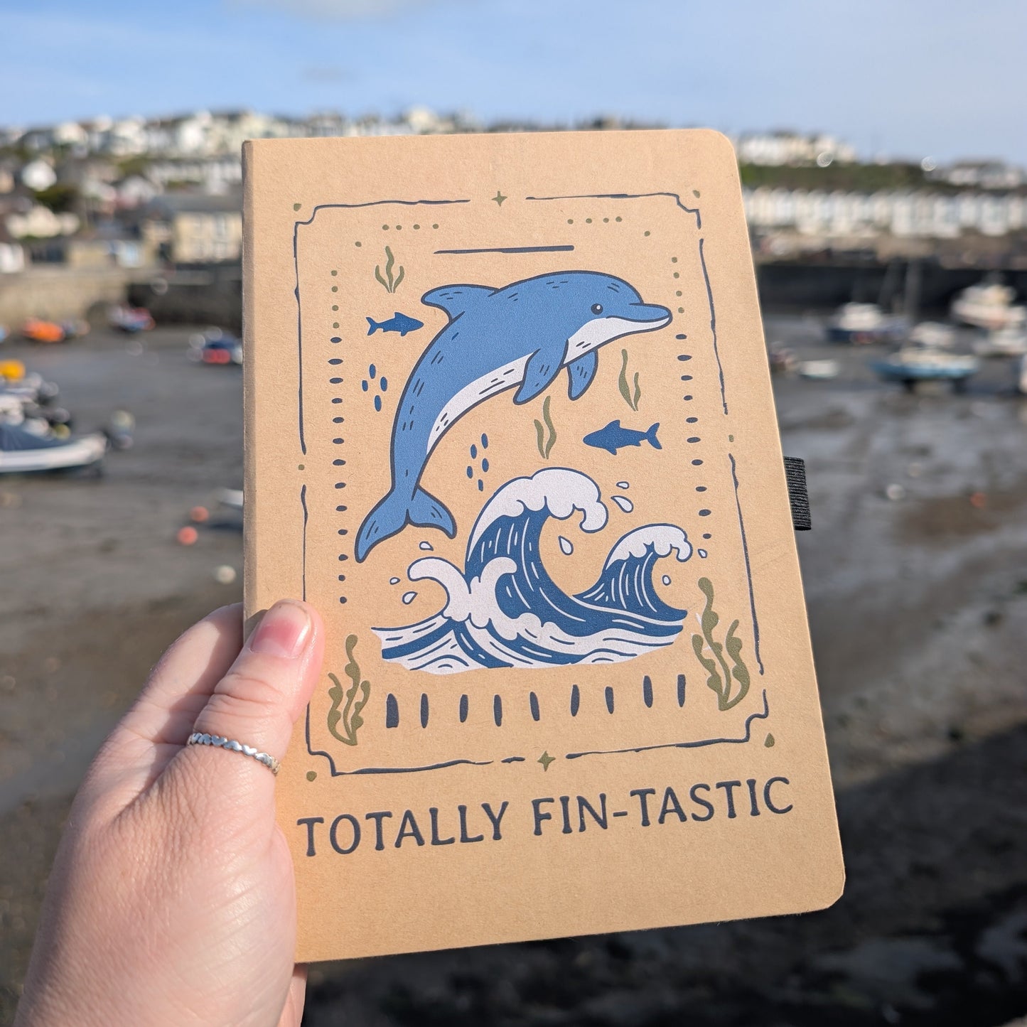 Notebook with dolphin design held by a hand, with a coastal background.