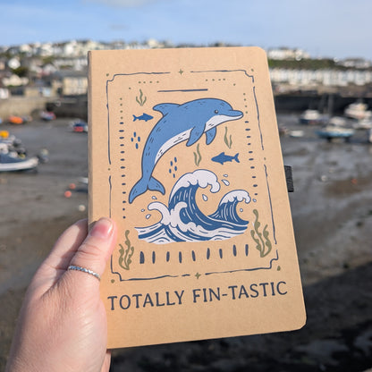 Notebook with dolphin design held by a hand, with a coastal background.