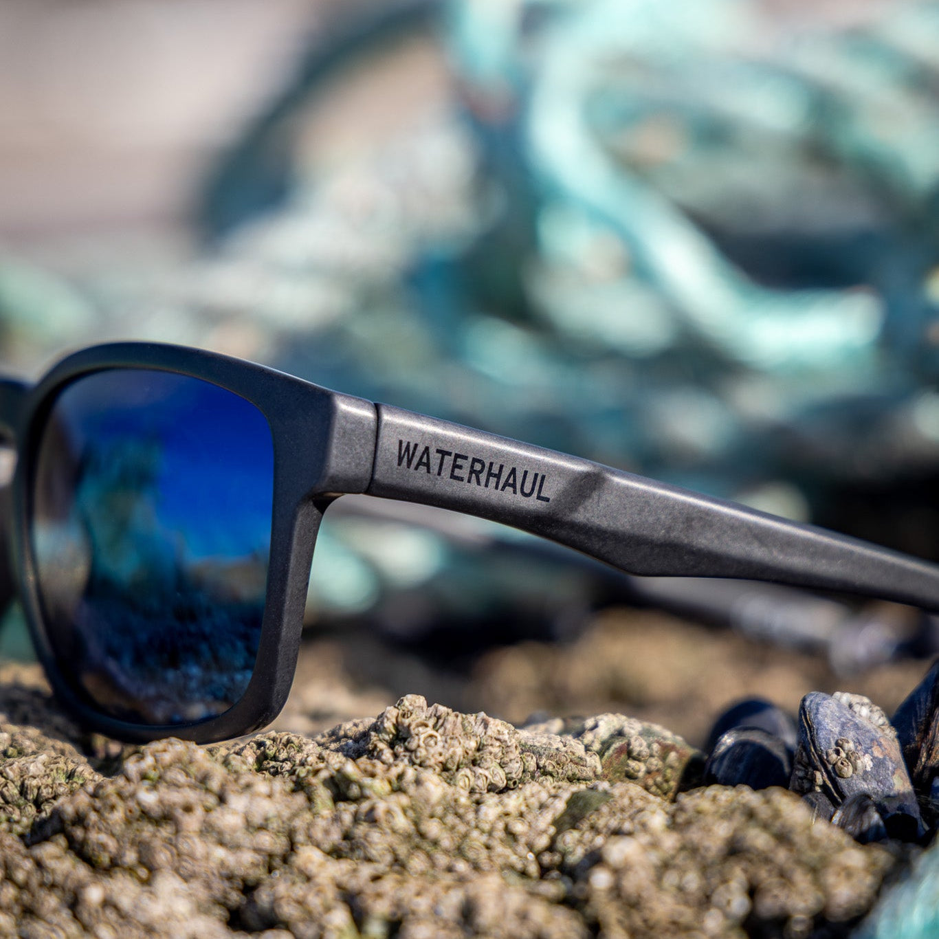 Towan Black Blue Mirror Polarised Lens Sunglasses on a rock with ghost fishing nets in the background. 