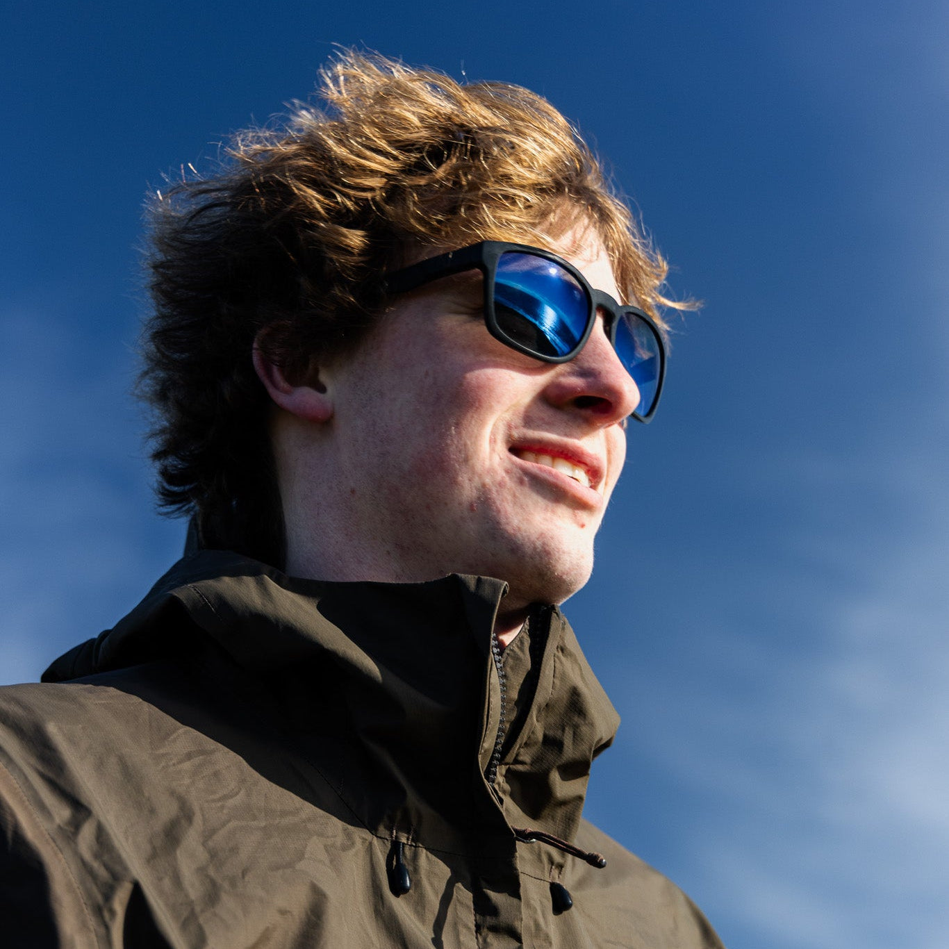 Person wearing black sunglasses with a blue mirror polarised lens against a blue sky.