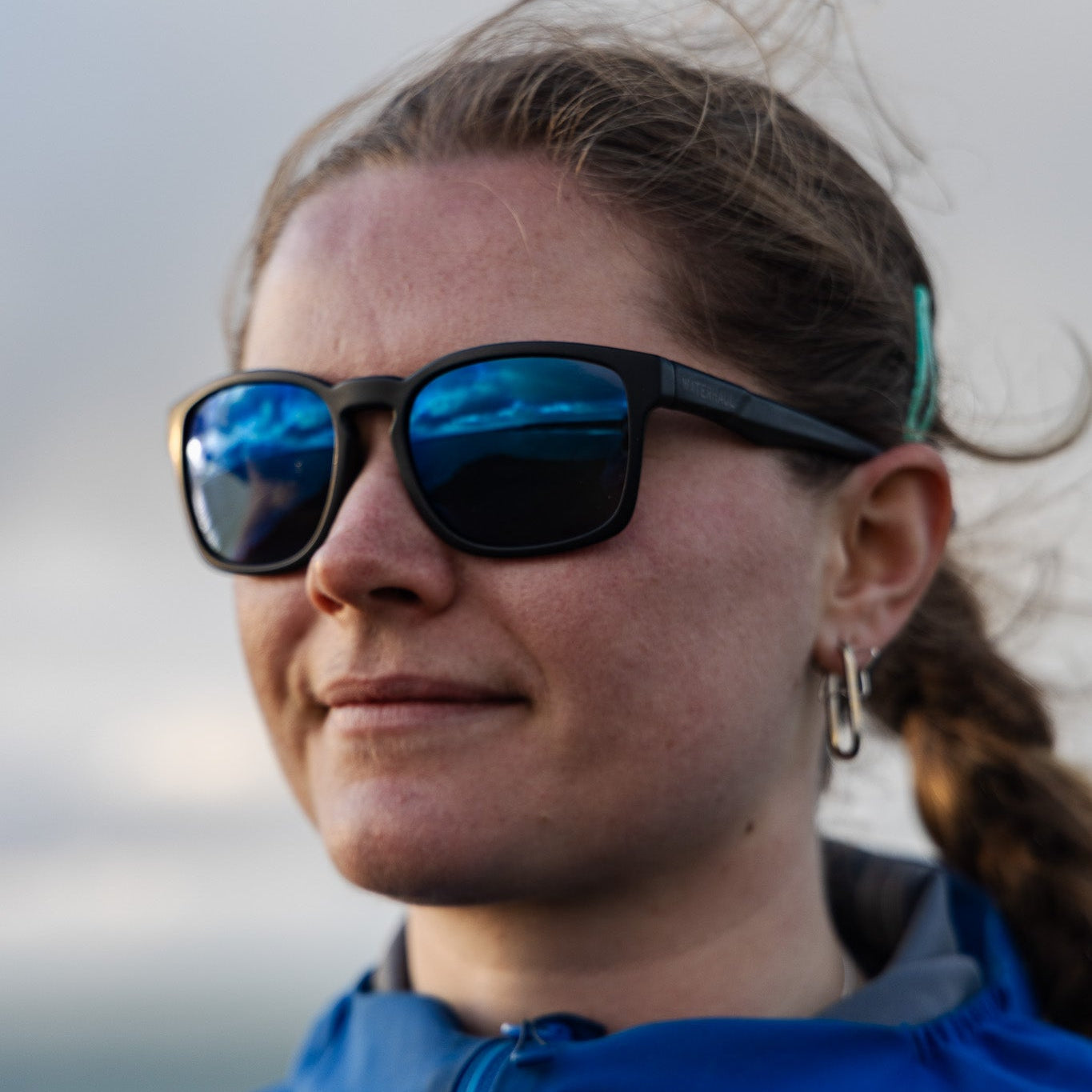 Person wearing black recycled fishing net sunglasses with a blue reflective lens in front of a blurred background