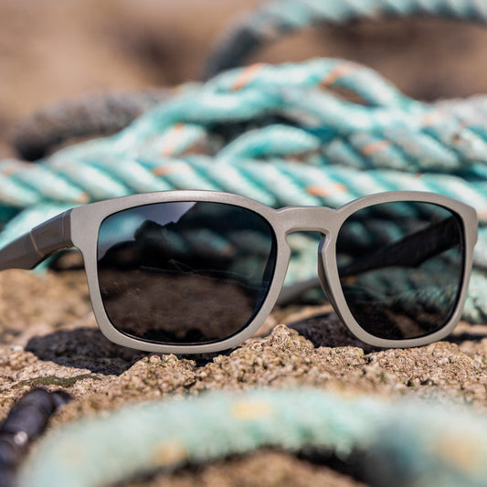Green-grey square sunglasses with dark lenses resting on a sandy surface with a coiled turquoise rope in the blurred background.