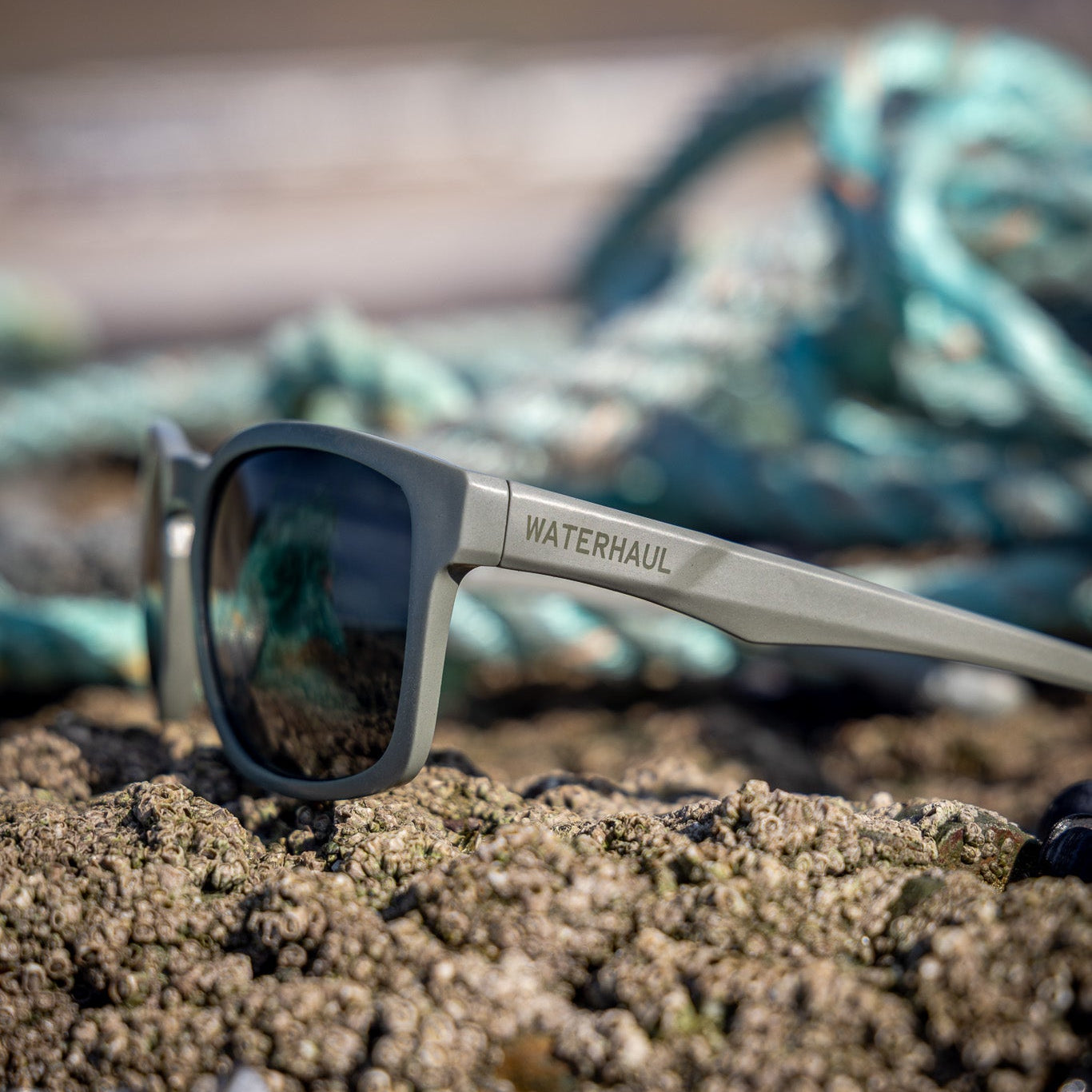 A pair of WATERHAUL sunglasses with a matte gray frame and dark lenses, resting on a sandy beach with shells in the foreground and a blurred blue-green rope in the background.