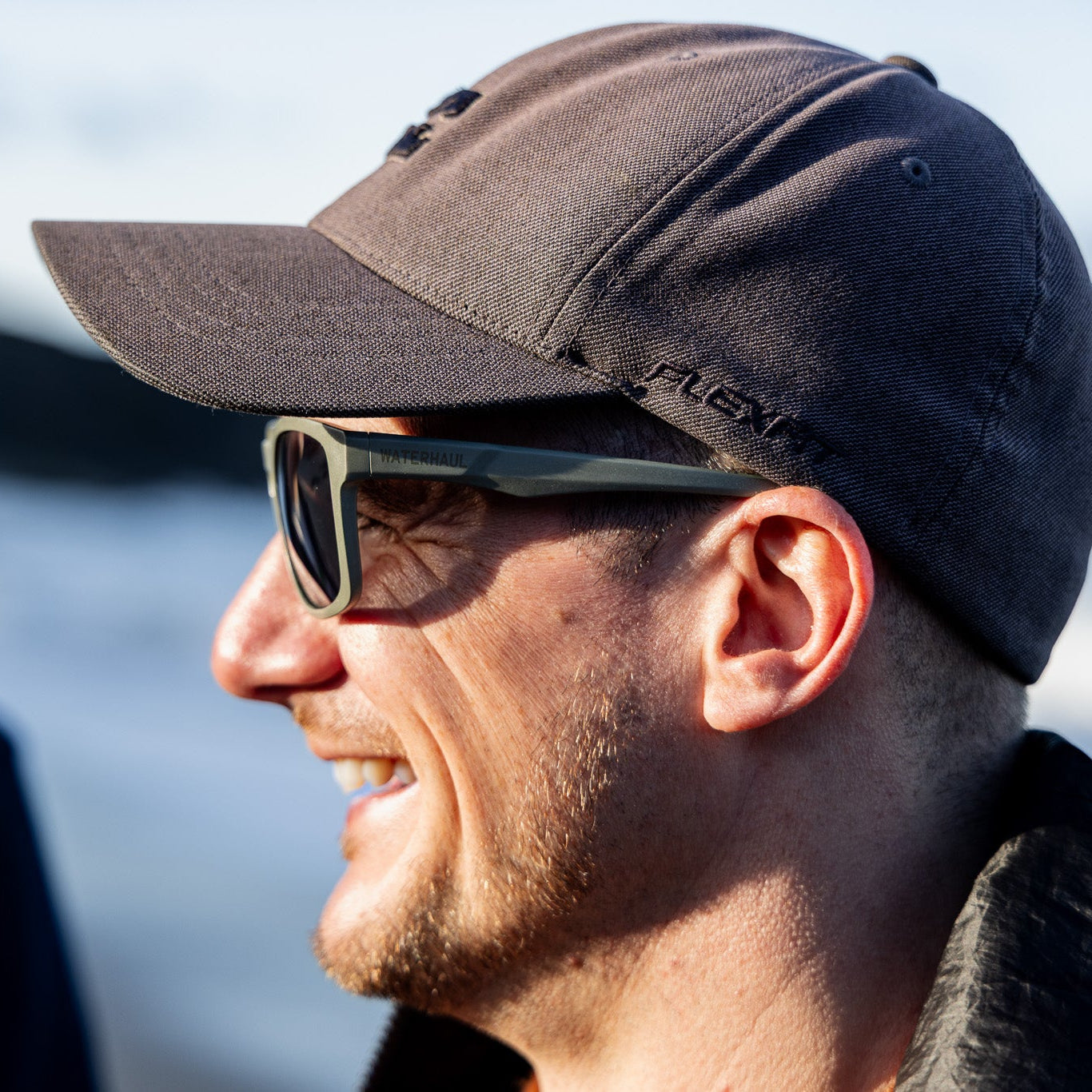 A man in side profile wearing green recycled fishing net sunglasses, with a blurred outdoor background.