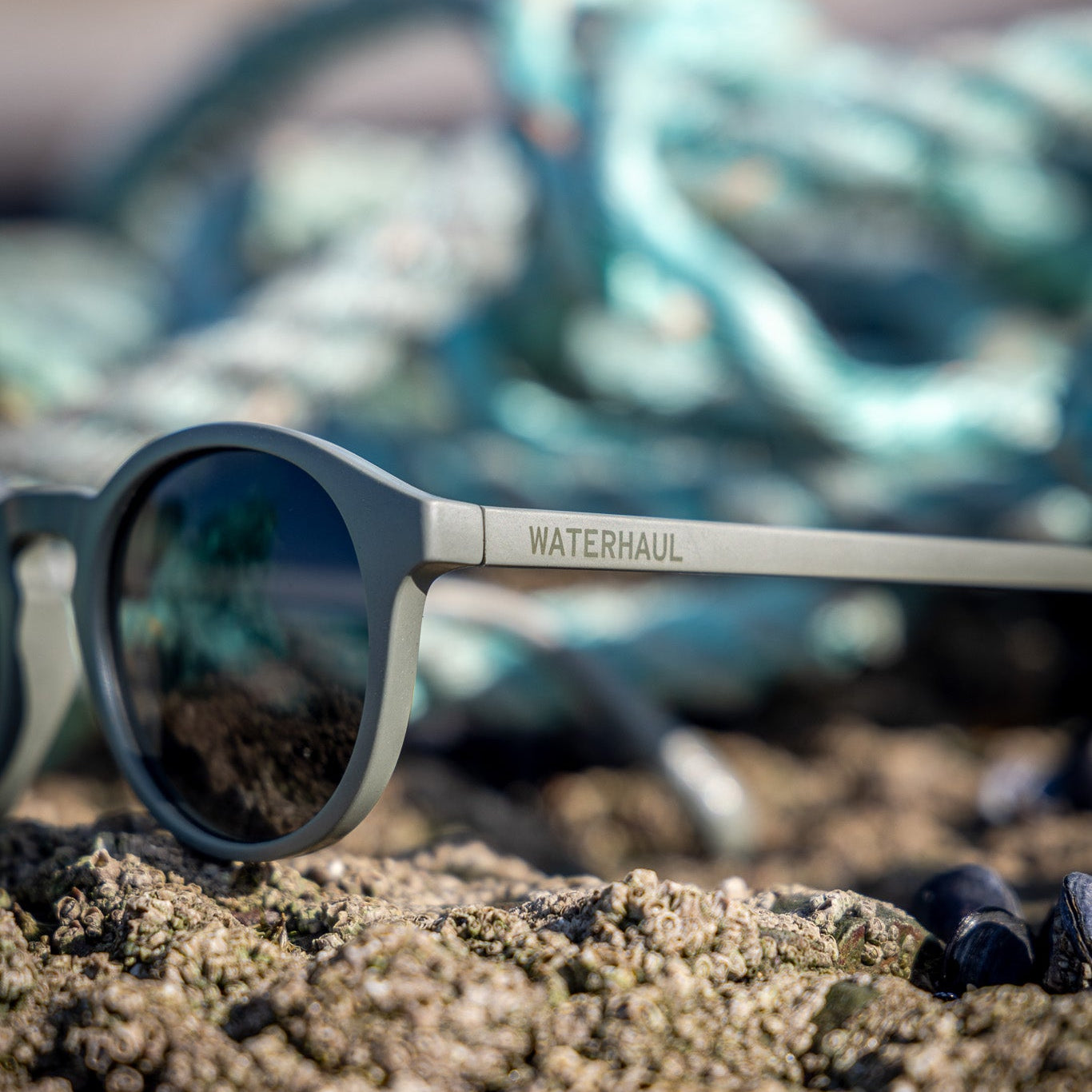 Waterhaul Trevellas Raw ocean plastic sunglasses on a sandy surface with fishing nets in the background