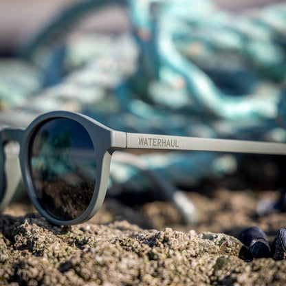 Waterhaul Trevellas Raw ocean plastic sunglasses on a sandy surface with fishing nets in the background