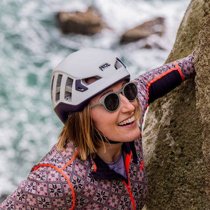 A woman wearing a white climbing helmet, recycled ocean waste sunglasses, and a patterned climbing jacket with orange accents is climbing a large rock face next to the ocean, with waves visible in the background.