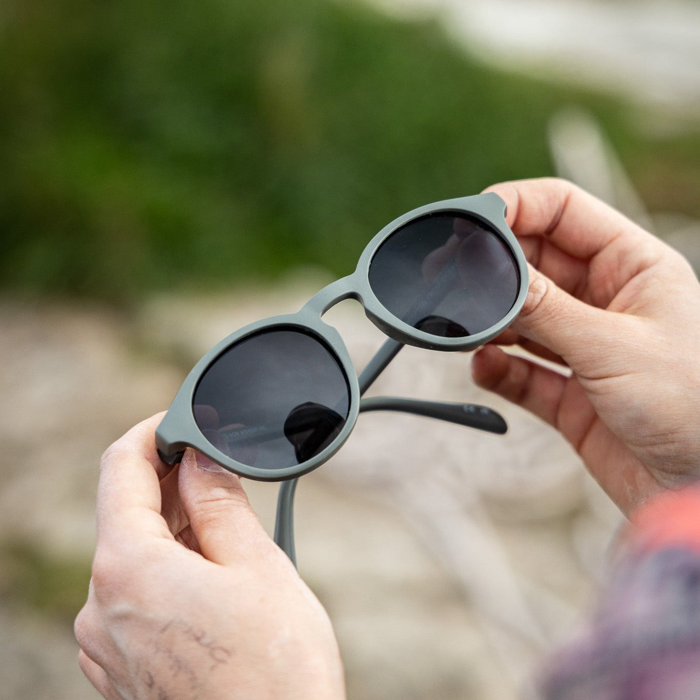 Person holding a pair of green-grey polarised sunglasses with a blurred natural background