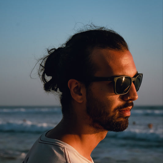 Man with recycled ocean plastic sunglasses and a bun standing on a beach with ocean waves in the background
