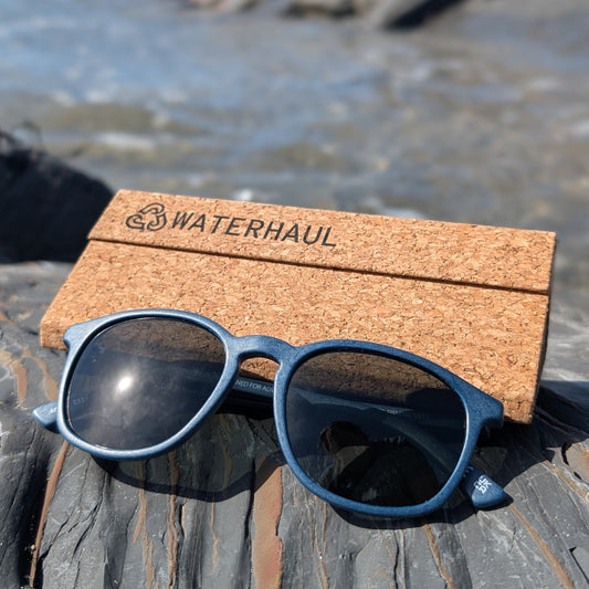 Blue sunglasses with a cork case labeled 'WATERHAUL' on a rocky surface near water.