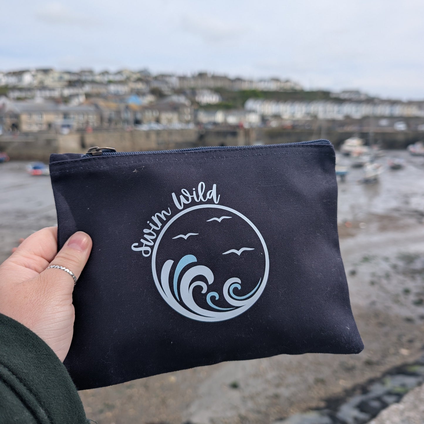 Navy pouch with 'Swim Wild' branding held by a hand, with a coastal background.