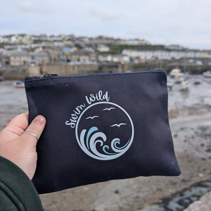 Navy pouch with 'Swim Wild' branding held by a hand, with a coastal background.