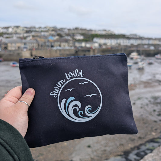 Navy pouch with 'Swim Wild' branding held by a hand, with a coastal background.