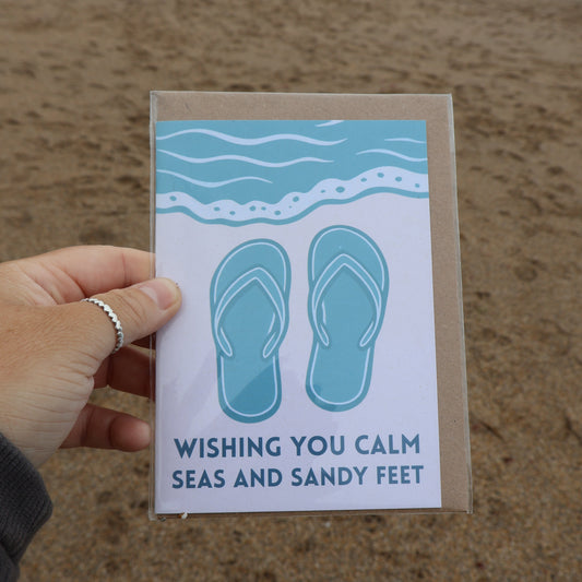 Hand holding a recycled greeting card with flip-flop design and text at the beach. 
