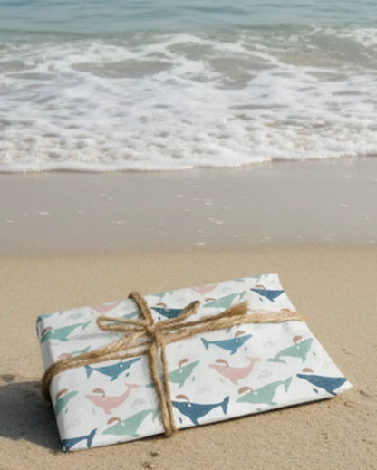 Festive whales wearing santa hats wrapping paper on a sandy beach with ocean waves in the background